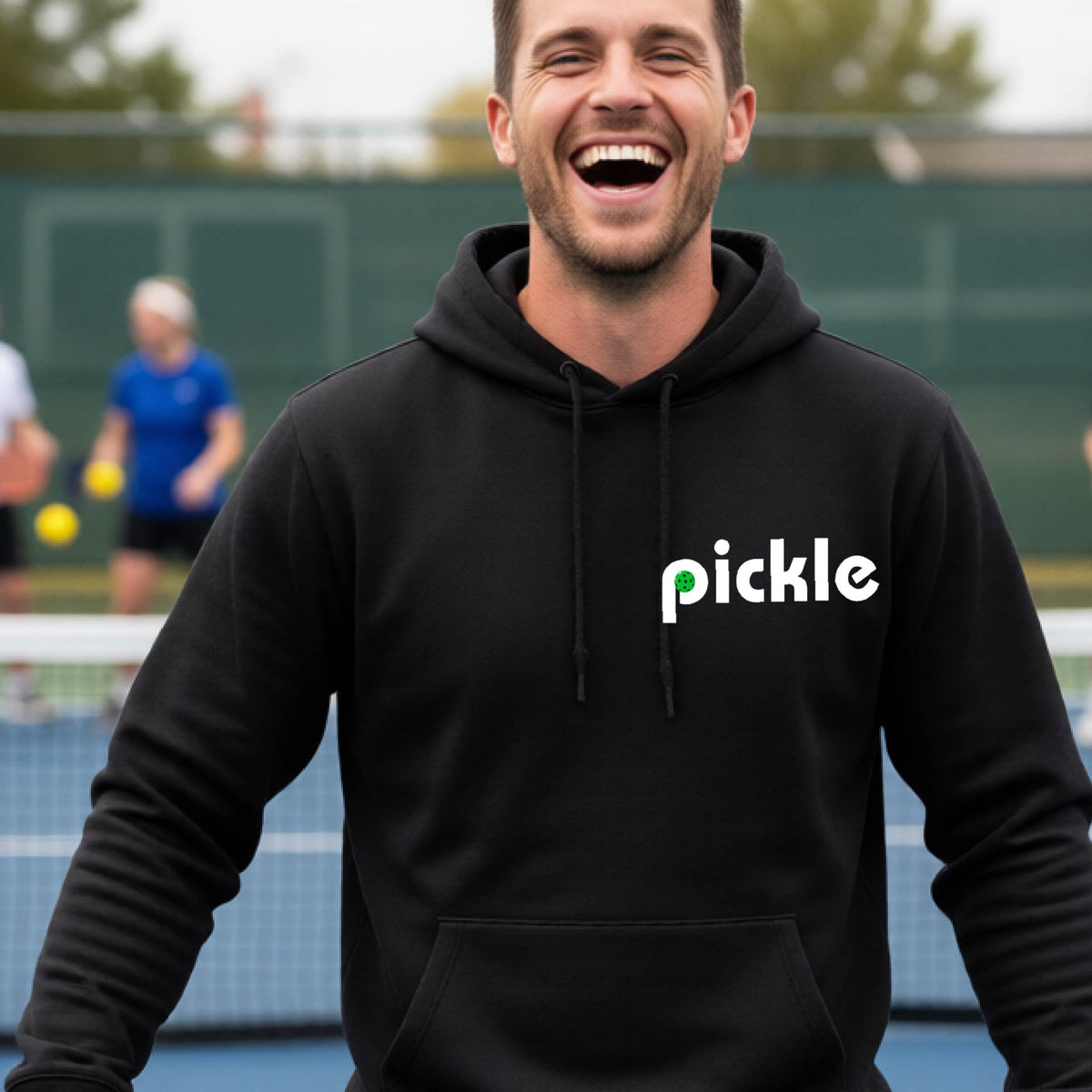 A close-up of a black unisex hoodie pullover with a white graphic of ‘PICKLE’ with a green pickleball integrated into the letter P printed on the front, in front of a pickleball court. 
