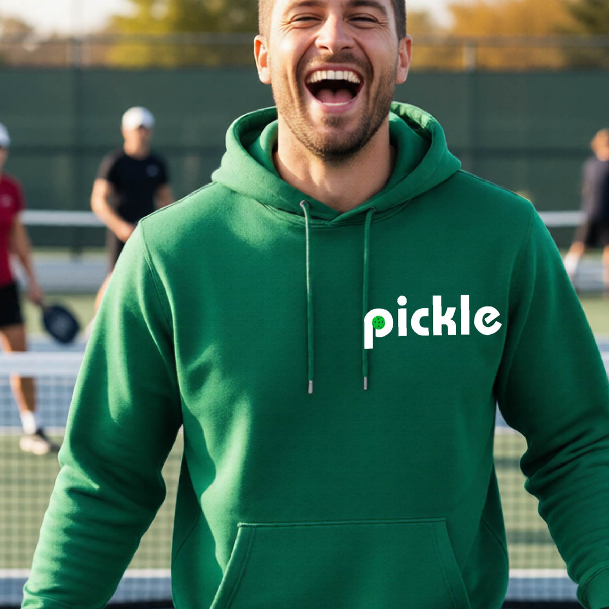 A close-up of a green unisex hoodie pullover with a white graphic of ‘PICKLE’ with a green pickleball integrated into the letter P printed on the front, in front of a pickleball court. 