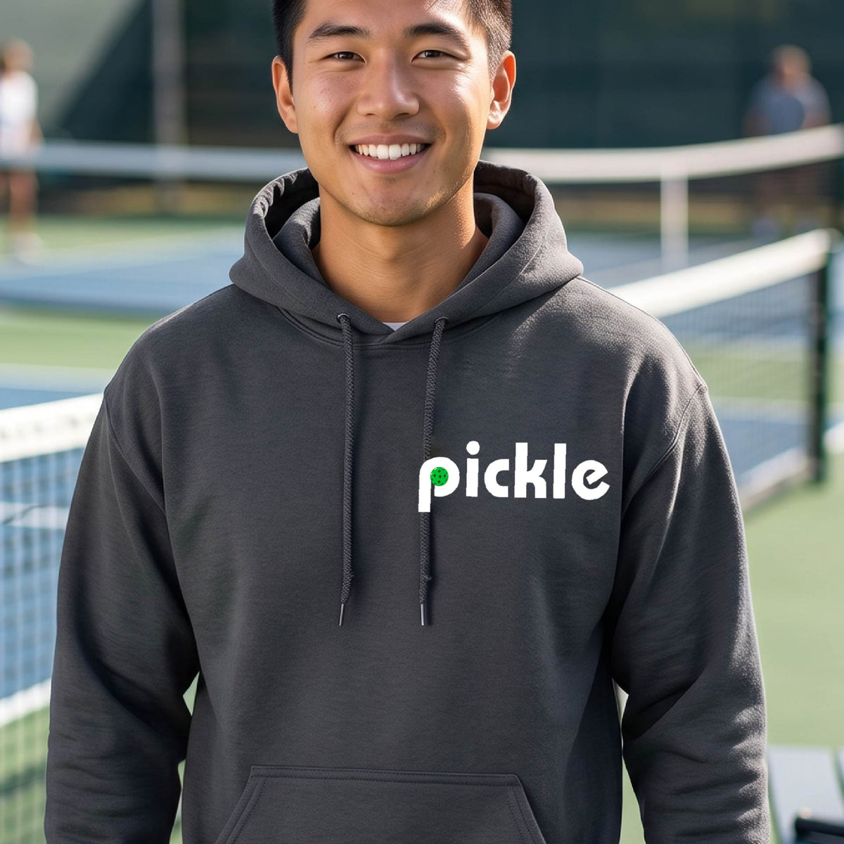 A close-up of a dark gray unisex hoodie pullover with a white graphic of ‘PICKLE’ with a green pickleball integrated into the letter P printed on the front, in front of a pickleball court. 