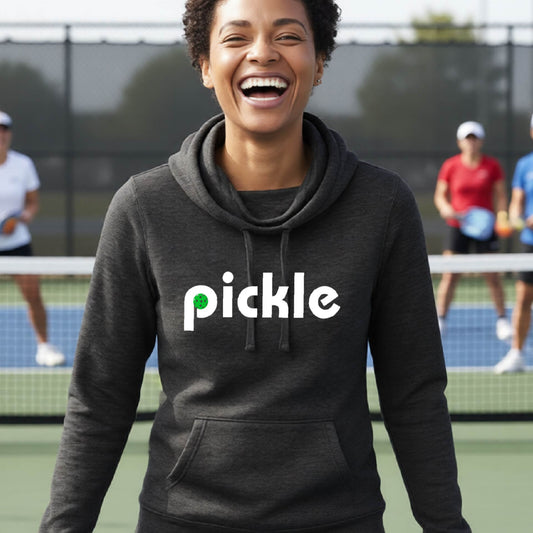 A close-up of a woman wearing a black cowl-neck hoodie pullover with a white graphic of ‘PICKLE’ with a green pickleball integrated into the letter P printed on the front, in front of a pickleball court. 