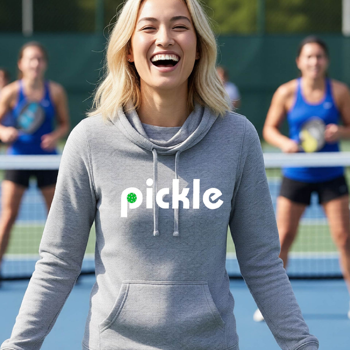 A close-up of a woman wearing a light gray cowl-neck hoodie pullover with a white graphic of ‘PICKLE’ with a green pickleball integrated into the letter P printed on the front, in front of a pickleball court. 