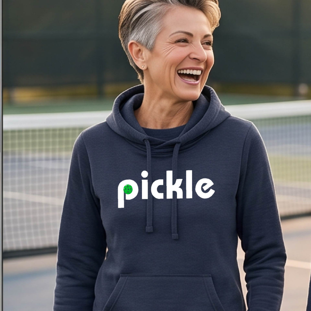A close-up of a woman wearing a navy blue cowl-neck hoodie pullover with a white graphic of ‘PICKLE’ with a green pickleball integrated into the letter P printed on the front, in front of a pickleball court. 