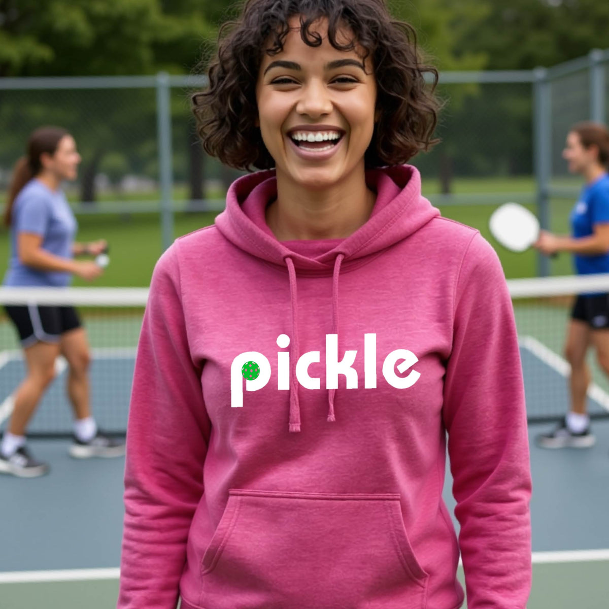 A close-up of a woman wearing a pink cowl-neck hoodie pullover with a white graphic of ‘PICKLE’ with a green pickleball integrated into the letter P printed on the front, in front of a pickleball court. 