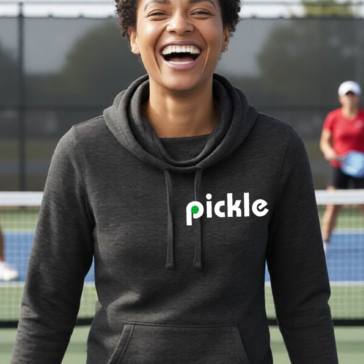 A close-up of a woman wearing a black cowl-neck hoodie pullover with a white graphic of ‘PICKLE’ with a green pickleball integrated into the letter P printed on the front, in front of a pickleball court. 