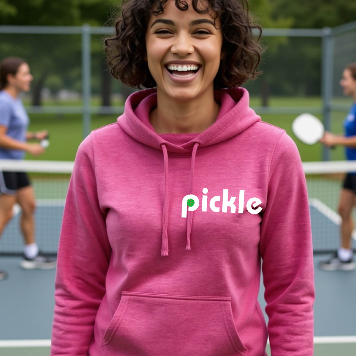 A close-up of a woman wearing a pink cowl-neck hoodie pullover with a white graphic of ‘PICKLE’ with a green pickleball integrated into the letter P printed on the front, in front of a pickleball court. 