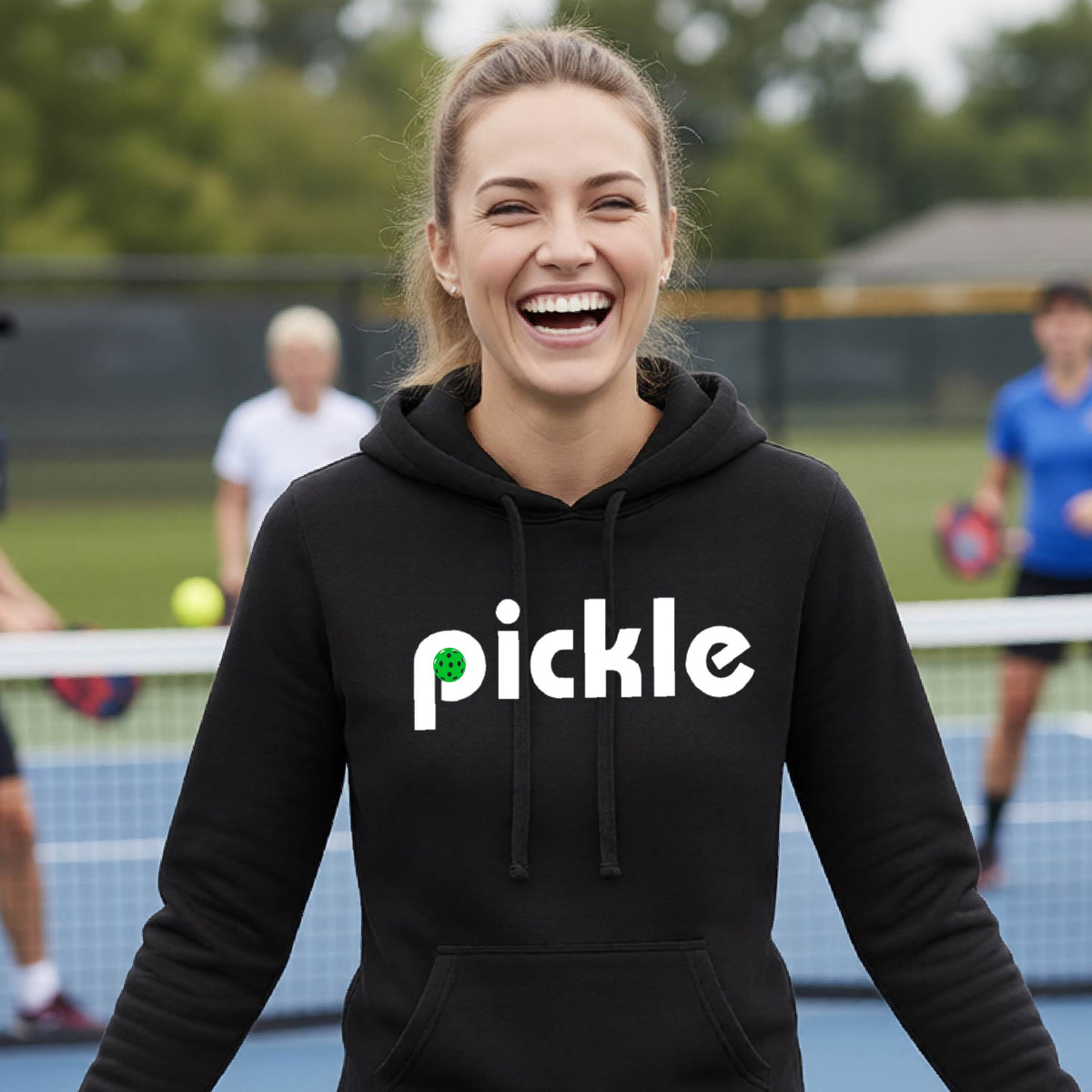 A smiling woman with blonde hair stands on a pickleball court wearing a black hoodie with the word 'pickle' printed across the chest in white, with a small green pickle graphic inside the letter 'iP. Two blurred players are visible in the background on the court.