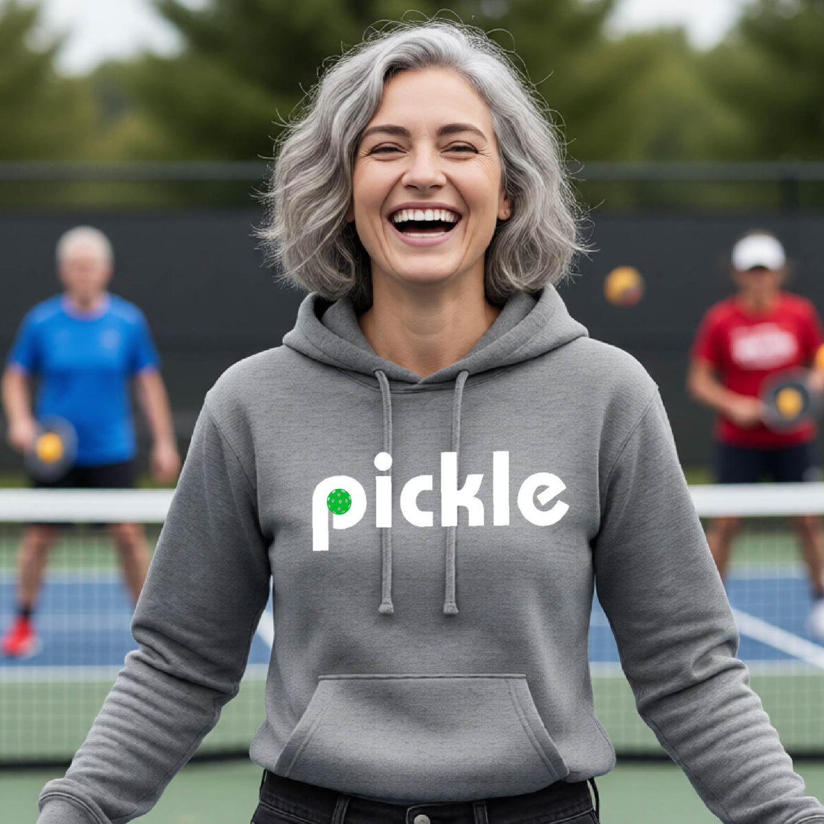 A smiling woman with gray hair stands on a pickleball court wearing a gray hoodie with the word 'pickle' printed across the chest in white, with a small green pickle graphic inside the letter 'iP. Two blurred players are visible in the background on the court.