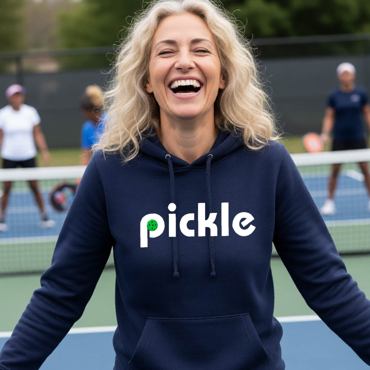 A smiling woman with blonde hair stands on a pickleball court wearing a navy blue hoodie with the word 'pickle' printed across the chest in white, with a small green pickle graphic inside the letter 'iP. Two blurred players are visible in the background on the court.