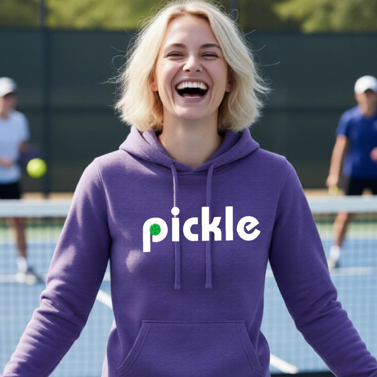 A smiling woman with blonde hair stands on a pickleball court wearing a purple hoodie with the word 'pickle' printed across the chest in white, with a small green pickle graphic inside the letter 'iP. Two blurred players are visible in the background on the court.