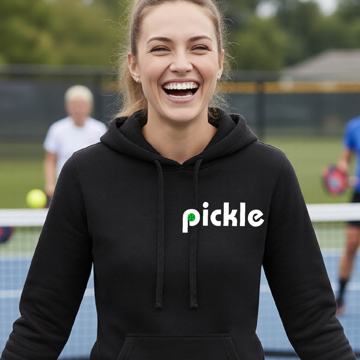 A smiling woman with blonde hair stands on a pickleball court wearing a black hoodie with the word 'pickle' printed across the chest in white, with a small green pickle graphic inside the letter 'iP. Two blurred players are visible in the background on the court.