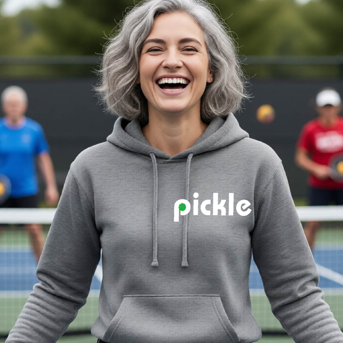 A smiling woman with gray hair stands on a pickleball court wearing a gray hoodie with the word 'pickle' printed across the chest in white, with a small green pickle graphic inside the letter 'iP. Two blurred players are visible in the background on the court.