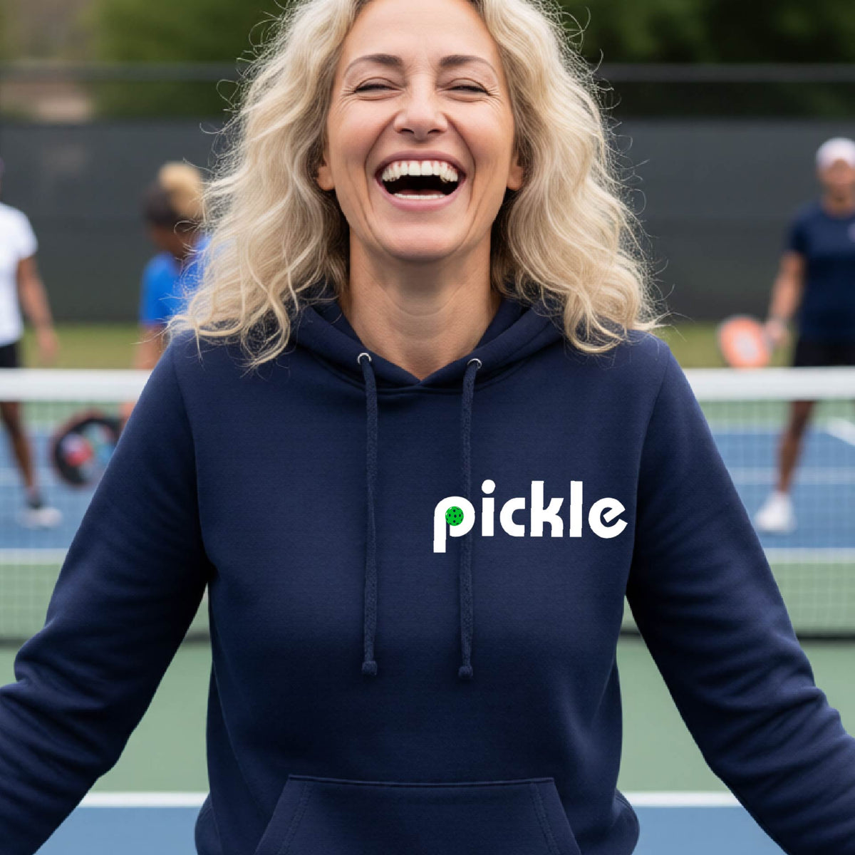 A smiling woman with blonde hair stands on a pickleball court wearing a navy blue hoodie with the word 'pickle' printed across the chest in white, with a small green pickle graphic inside the letter 'iP. Two blurred players are visible in the background on the court.
