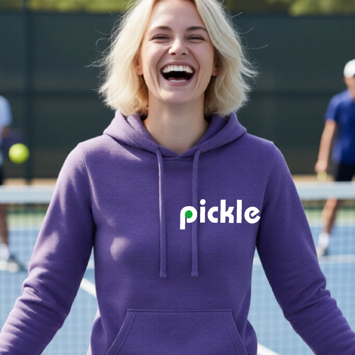 A smiling woman with blonde hair stands on a pickleball court wearing a purple hoodie with the word 'pickle' printed across the chest in white, with a small green pickle graphic inside the letter 'iP. Two blurred players are visible in the background on the court.
