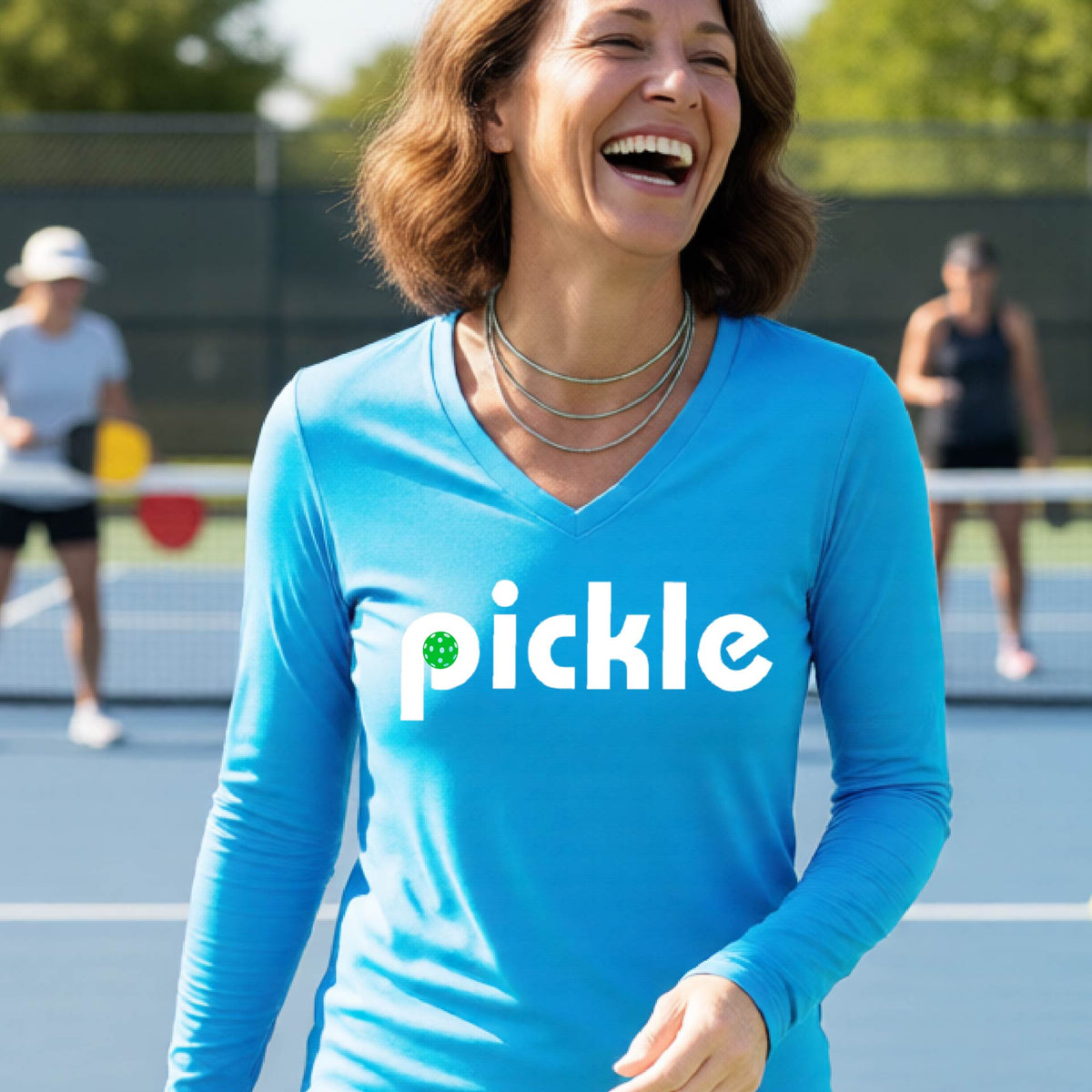 A woman wearing a bright blue long-sleeve v-neck t-shirt with a white graphic of ‘PICKLE’ with a green pickleball integrated into the letter P printed on the front, in front of a pickleball court. 