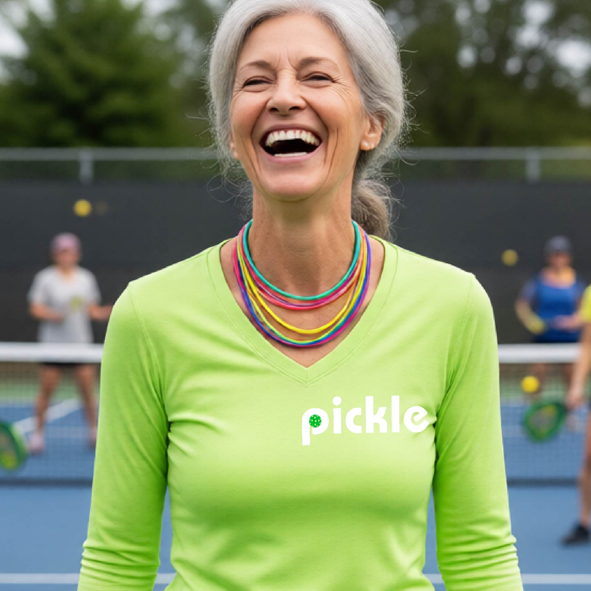 A woman wearing a lime green long-sleeve v-neck t-shirt with a white graphic of ‘PICKLE’ with a green pickleball integrated into the letter P printed on the front, in front of a pickleball court. 