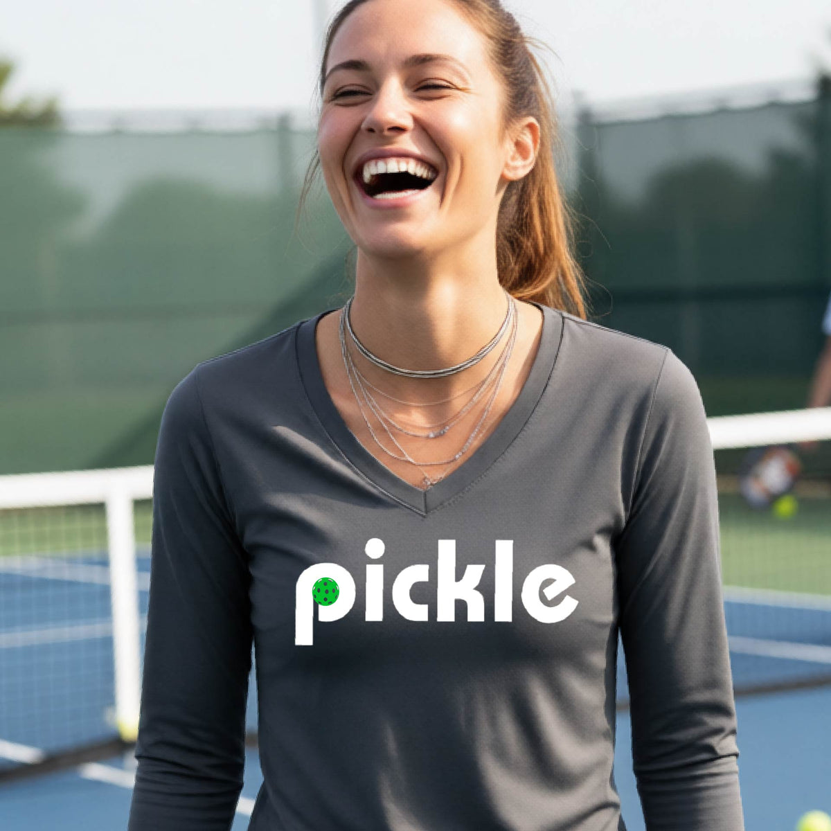 A woman wearing a dark gray long-sleeve v-neck t-shirt with a white graphic of ‘PICKLE’ with a green pickleball integrated into the letter P printed on the front, in front of a pickleball court. 