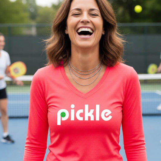A woman wearing a coral pink long-sleeve v-neck t-shirt with a white graphic of ‘PICKLE’ with a green pickleball integrated into the letter P printed on the front, in front of a pickleball court. 