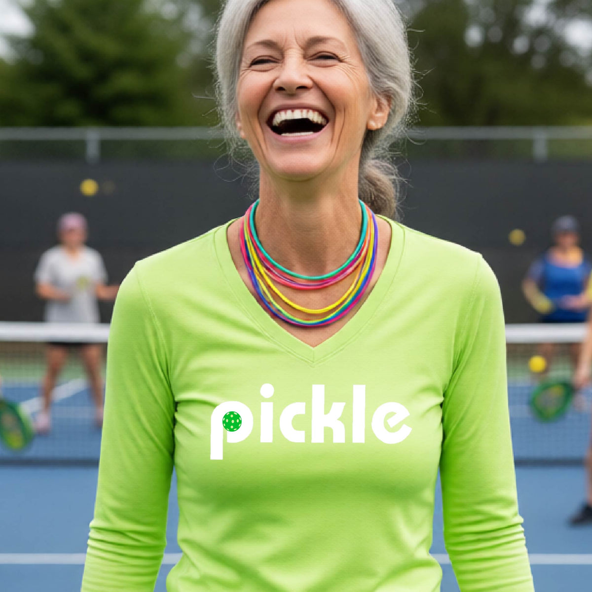 A woman wearing a lime green long-sleeve v-neck t-shirt with a white graphic of ‘PICKLE’ with a green pickleball integrated into the letter P printed on the front, in front of a pickleball court. 