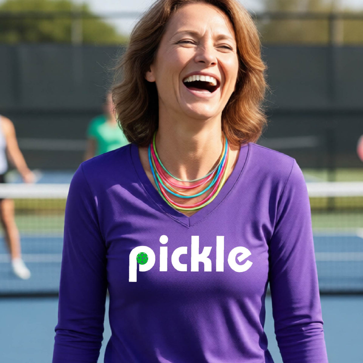A woman wearing a purple long-sleeve v-neck t-shirt with a white graphic of ‘PICKLE’ with a green pickleball integrated into the letter P printed on the front, in front of a pickleball court. 