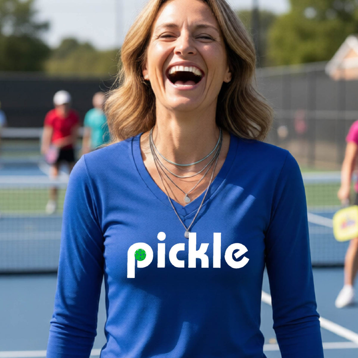 A woman wearing a royal blue long-sleeve v-neck t-shirt with a white graphic of ‘PICKLE’ with a green pickleball integrated into the letter P printed on the front, in front of a pickleball court. 