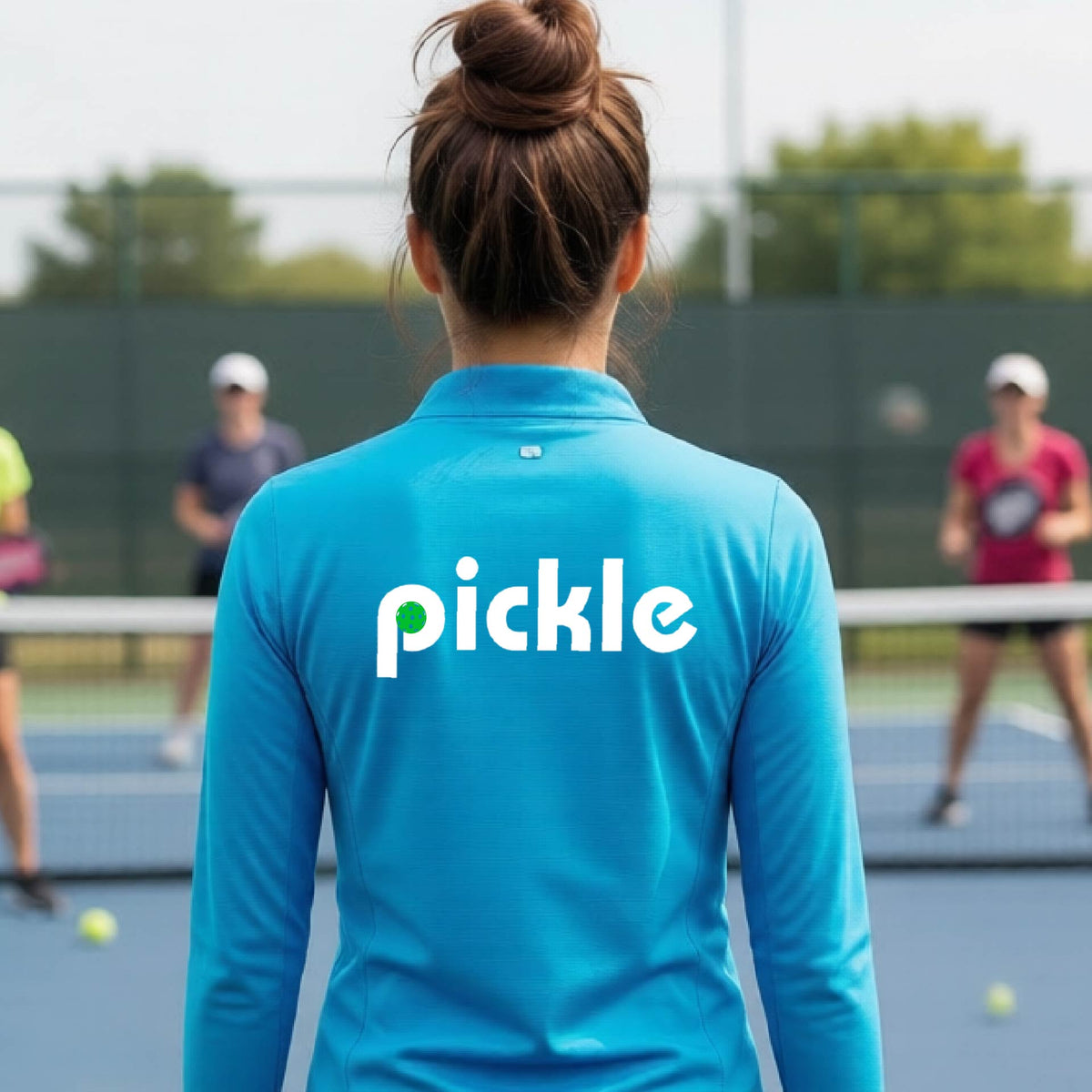 A woman wearing a blue quarter-zip shirt with a white graphic of ‘PICKLE’ with a green pickleball integrated into the letter P printed on the back, in front of a pickleball court. 
