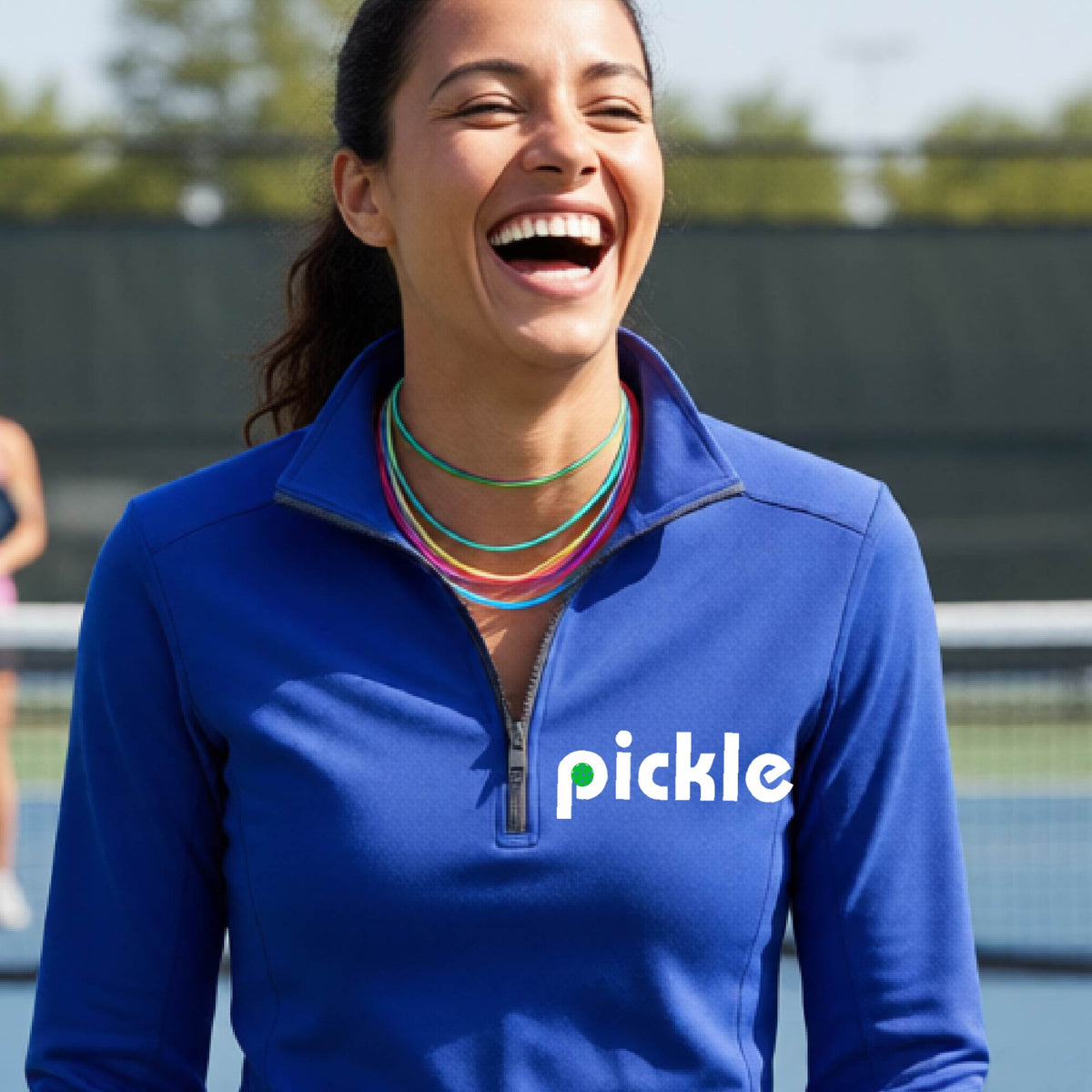A woman wearing a royal blue quarter-zip shirt with a white graphic of ‘PICKLE’ with a green pickleball integrated into the letter P printed on the front, in front of a pickleball court. 