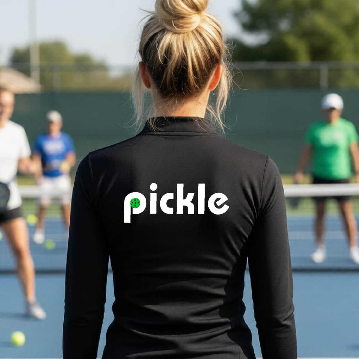 A woman wearing a black quarter-zip shirt with a white graphic of ‘PICKLE’ with a green pickleball integrated into the letter P printed on the back, in front of a pickleball court. 