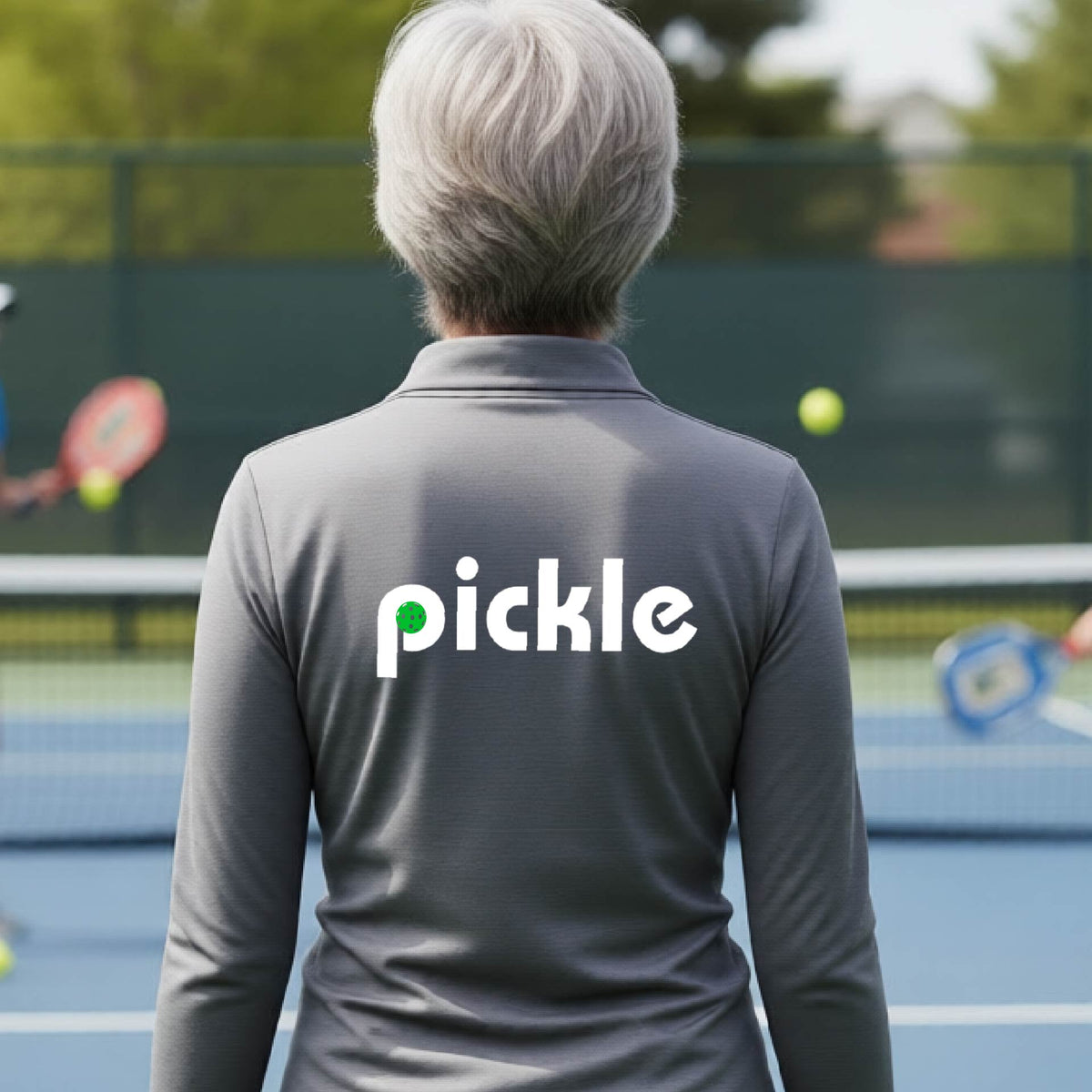 A woman wearing a gray quarter-zip shirt with a white graphic of ‘PICKLE’ with a green pickleball integrated into the letter P printed on the back, in front of a pickleball court. 