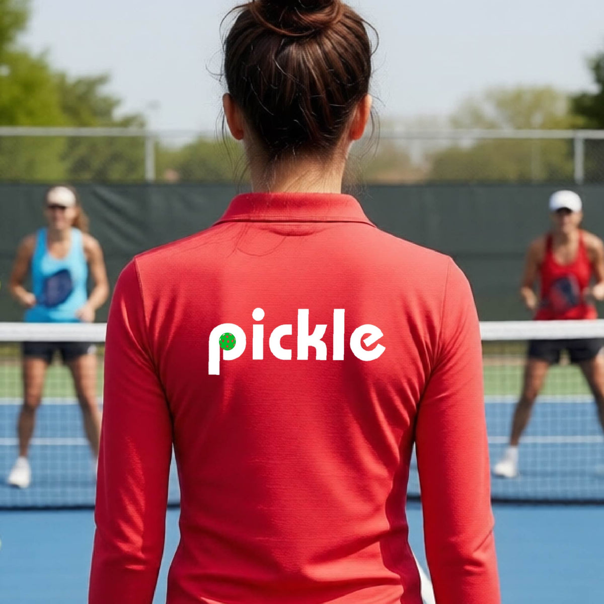 A woman wearing a red quarter-zip shirt with a white graphic of ‘PICKLE’ with a green pickleball integrated into the letter P printed on the back, in front of a pickleball court. 