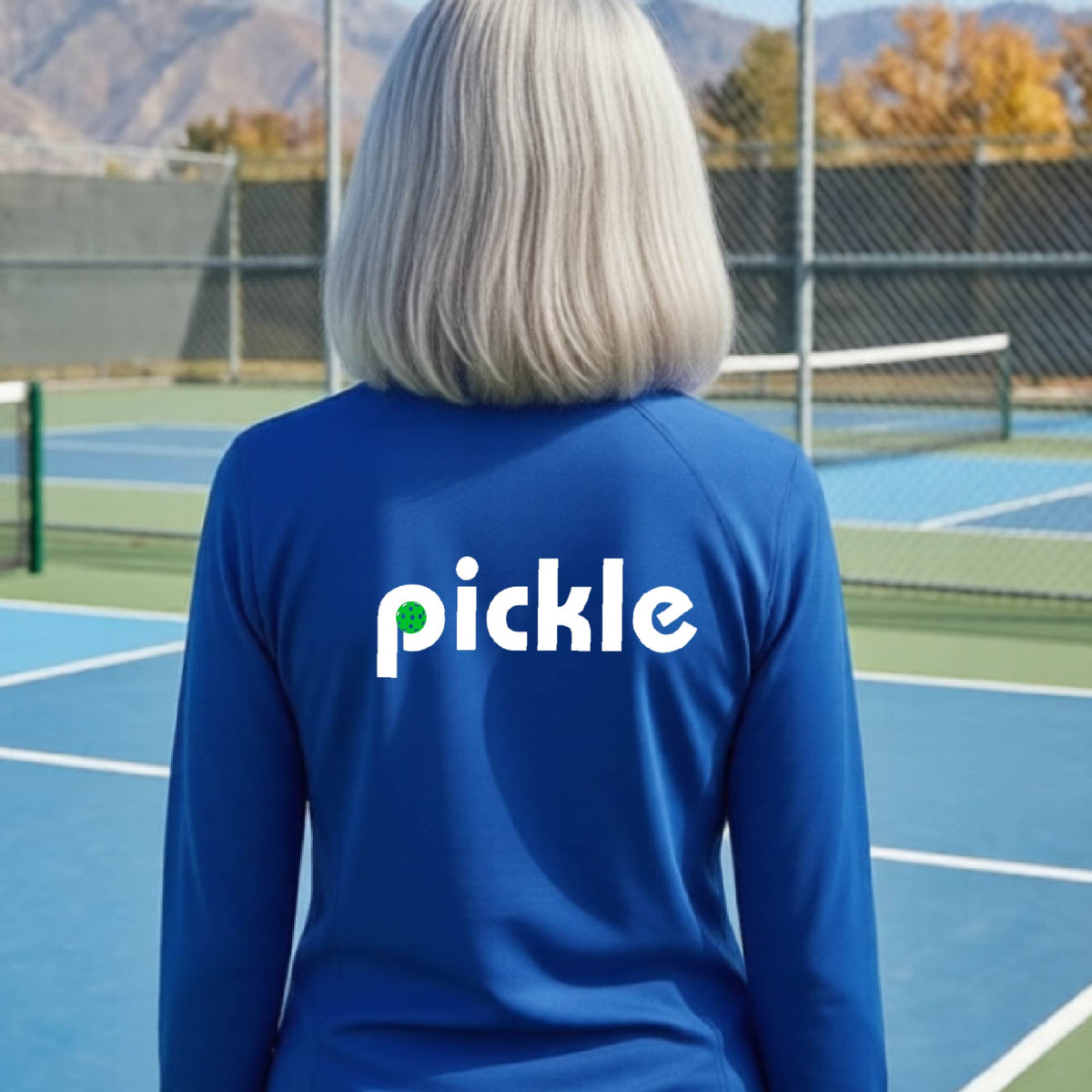 A woman wearing a roya bluel quarter-zip shirt with a white graphic of ‘PICKLE’ with a green pickleball integrated into the letter P printed on the back, in front of a pickleball court. 