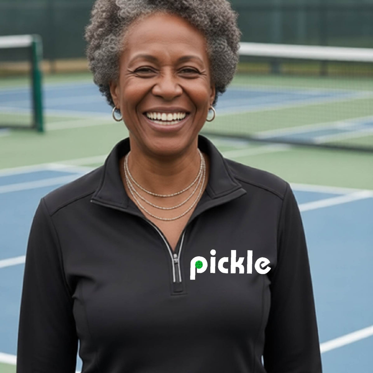 A woman wearing a black quarter-zip shirt with a white graphic of ‘PICKLE’ with a green pickleball integrated into the letter P printed on the front, in front of a pickleball court. 