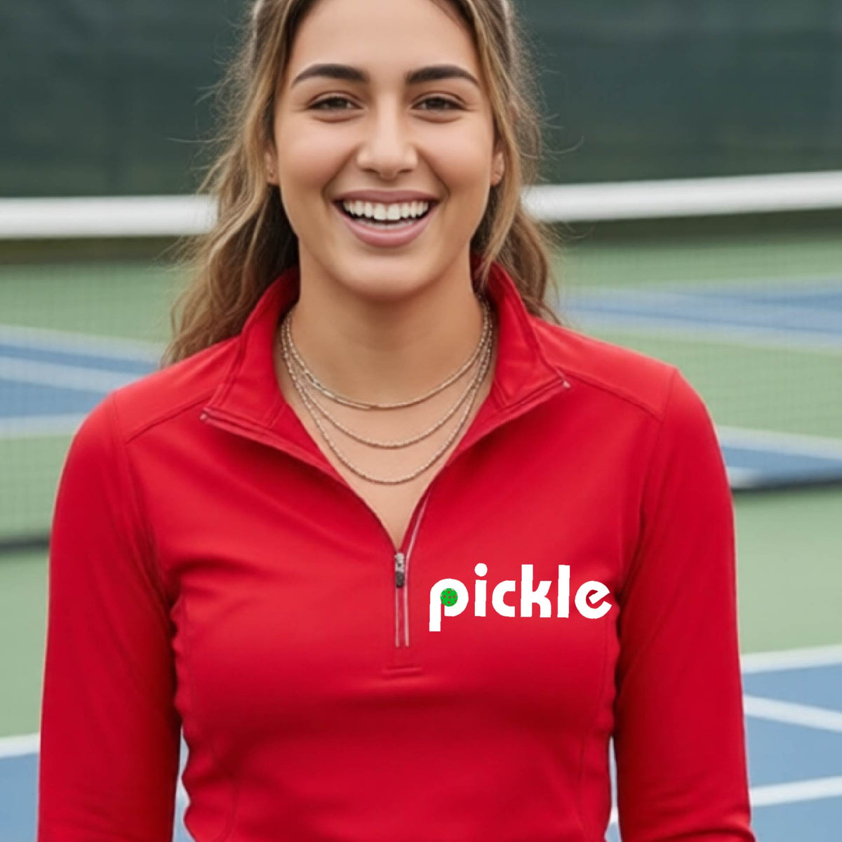 A woman wearing a red quarter-zip shirt with a white graphic of ‘PICKLE’ with a green pickleball integrated into the letter P printed on the front, in front of a pickleball court. 