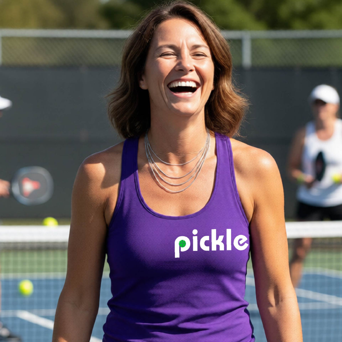 A woman laughing on a pickelball court wearing a purple athletic tank top with the word 'pickle' and a green pickelball icon inside the letter P, printed in white across the chest.