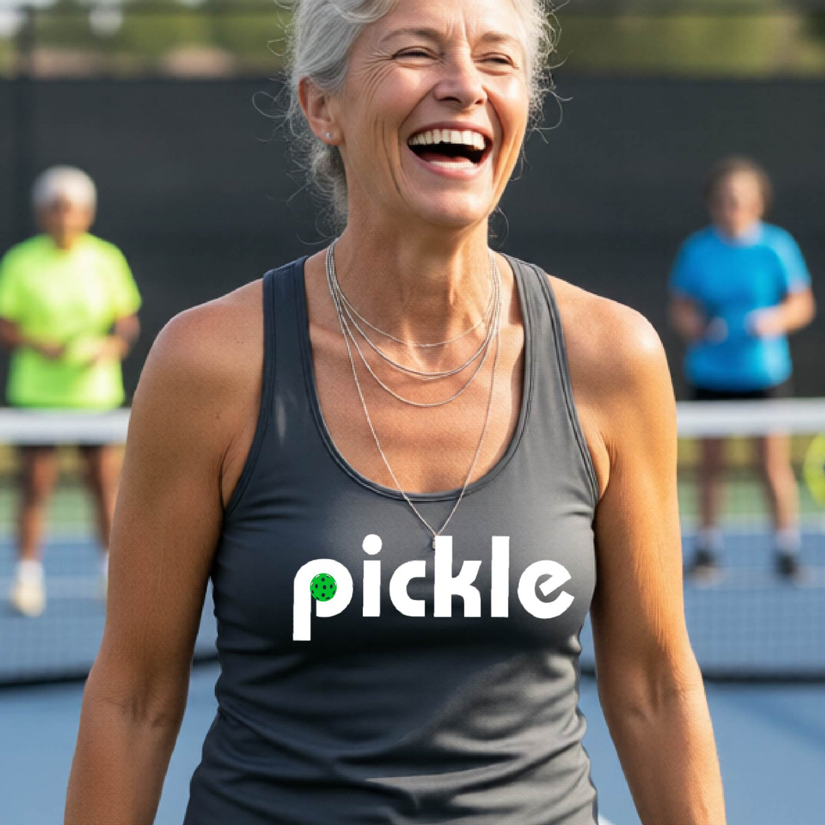 A woman laughing on a pickelball court wearing a dark gray athletic tank top with the word 'pickle' and a green pickelball icon inside the letter P, printed in white across the chest.
