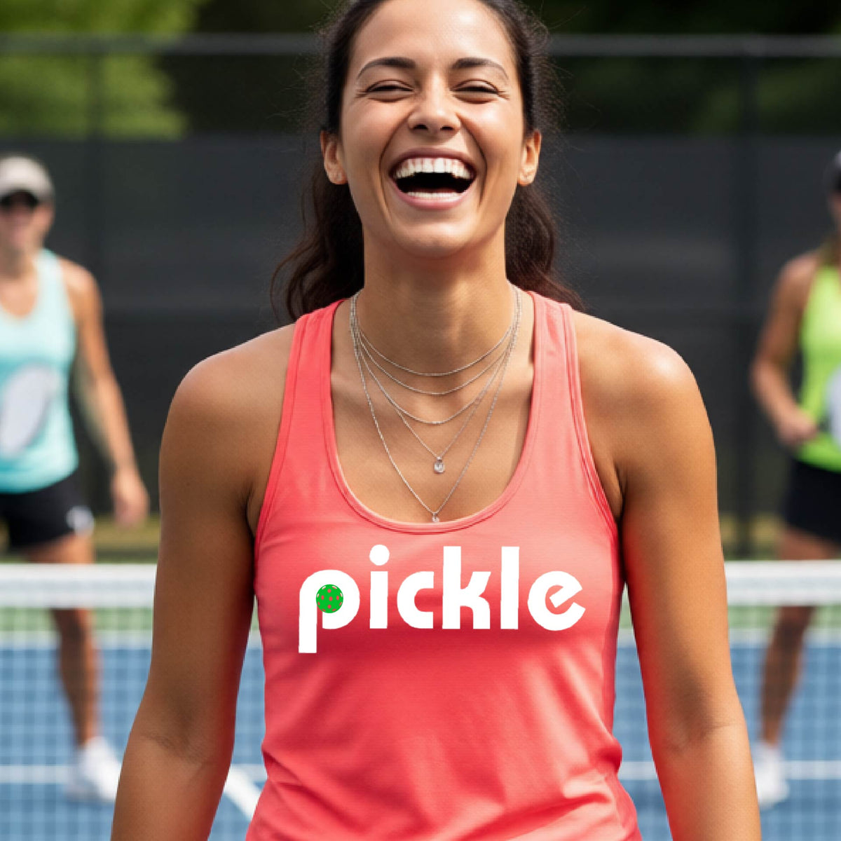 A woman laughing on a pickelball court wearing a coral pink athletic tank top with the word 'pickle' and a green pickelball icon inside the letter P, printed in white across the chest.