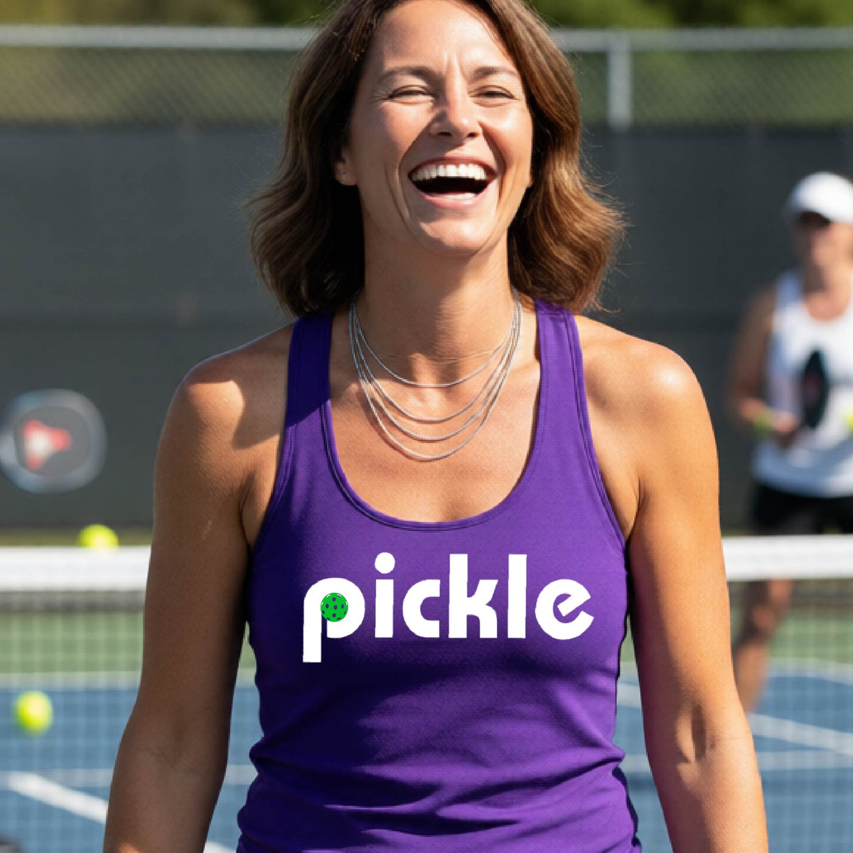 A woman laughing on a pickelball court wearing a purple athletic tank top with the word 'pickle' and a green pickelball icon inside the letter P, printed in white across the chest.