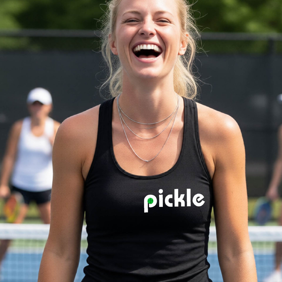 A woman laughing on a pickelball court wearing a black athletic tank top with the word 'pickle' and a green pickelball icon inside the letter P, printed in white across the chest.