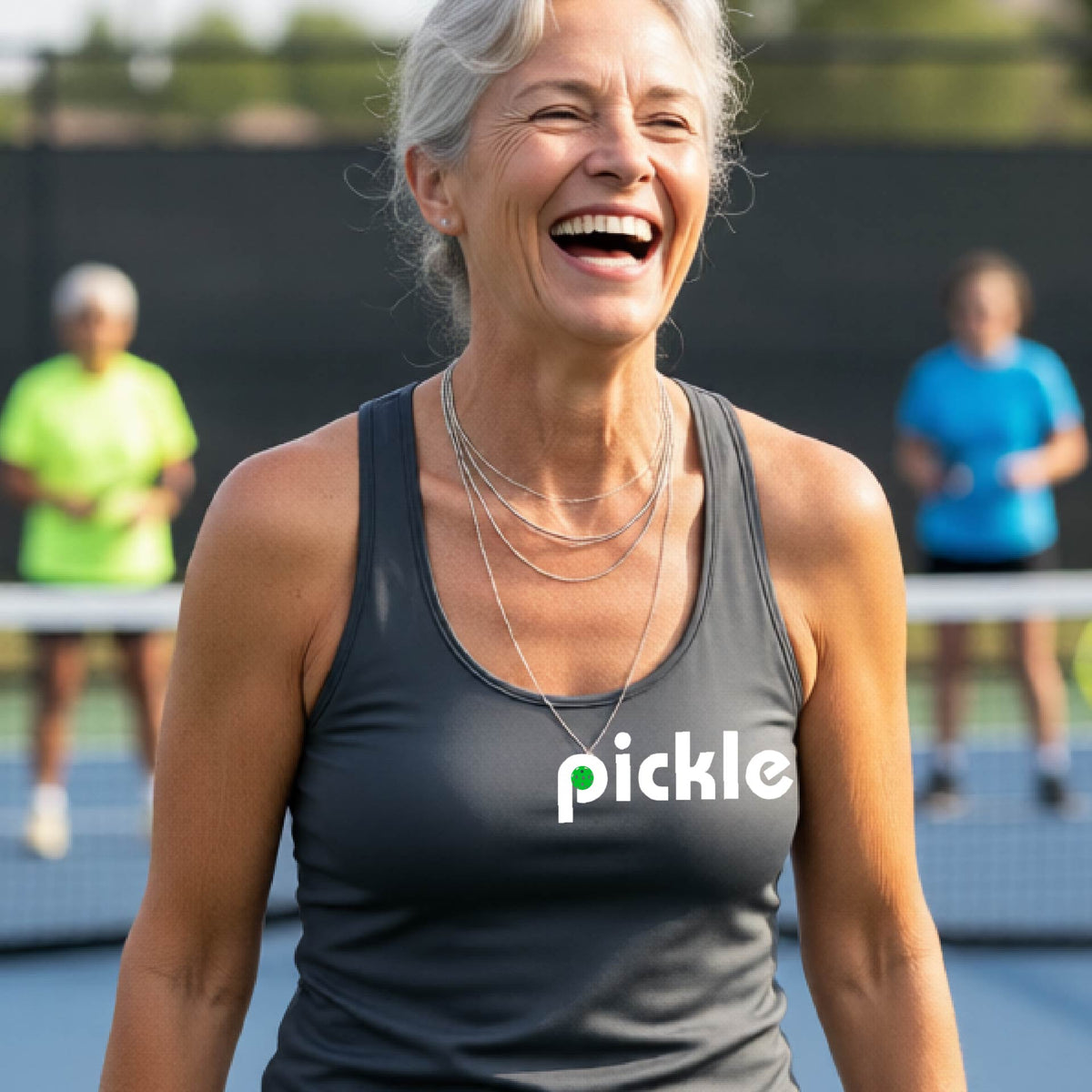 A woman laughing on a pickelball court wearing a dark gray athletic tank top with the word 'pickle' and a green pickelball icon inside the letter P, printed in white across the chest.