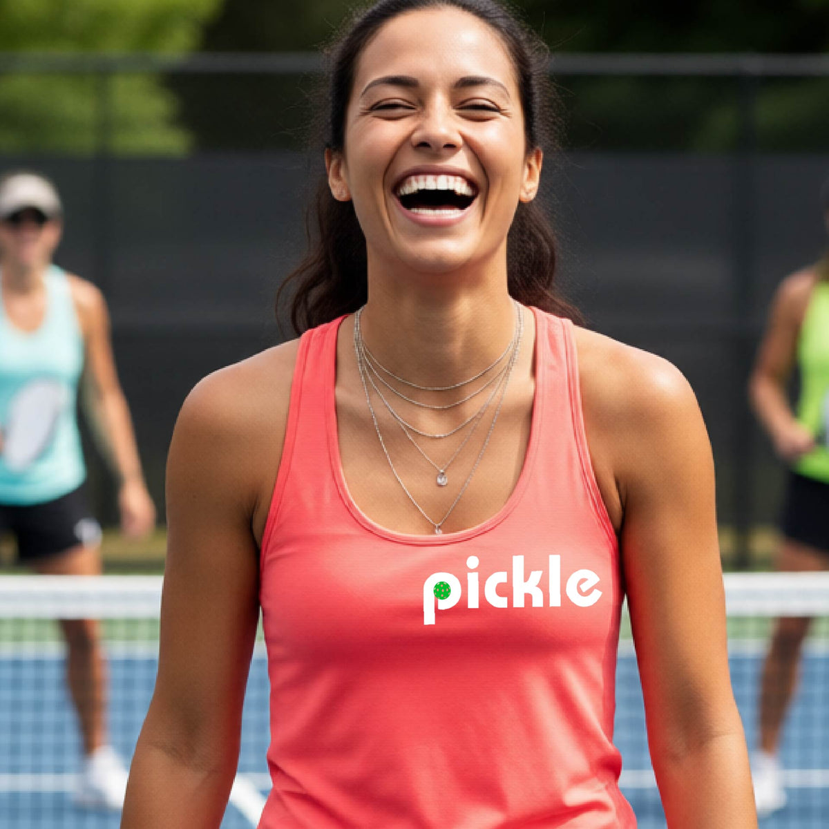 A woman laughing on a pickelball court wearing a coral pink athletic tank top with the word 'pickle' and a green pickelball icon inside the letter P, printed in white across the chest.