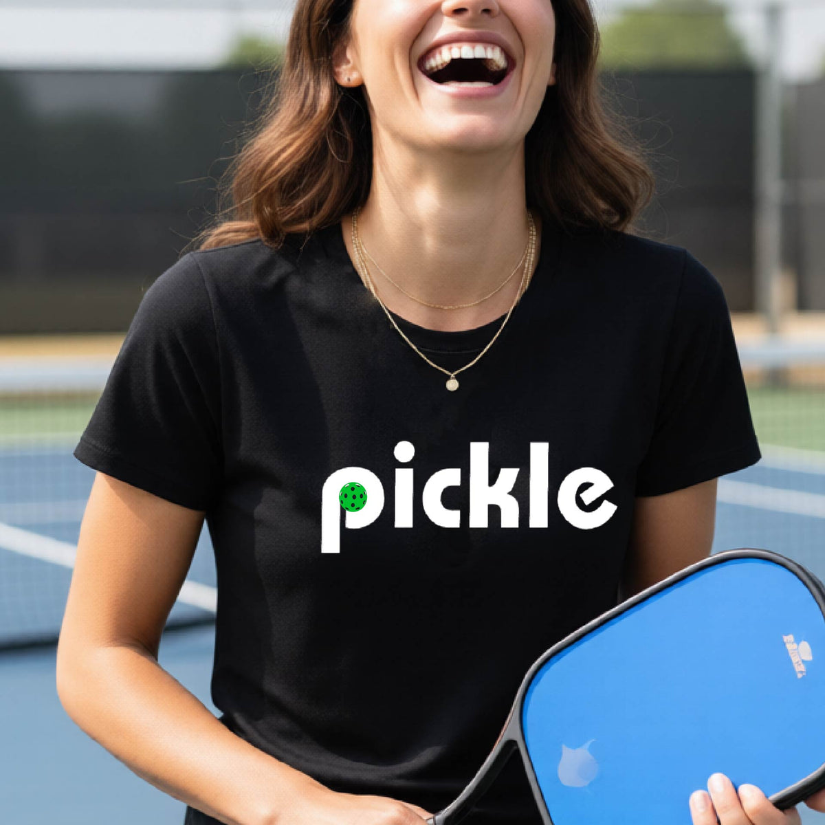 A woman wearing a black short-sleeve t-shirt with a white graphic of ‘PICKLE’ with a green pickleball integrated into the letter P printed on the front, in front of a pickleball court. 