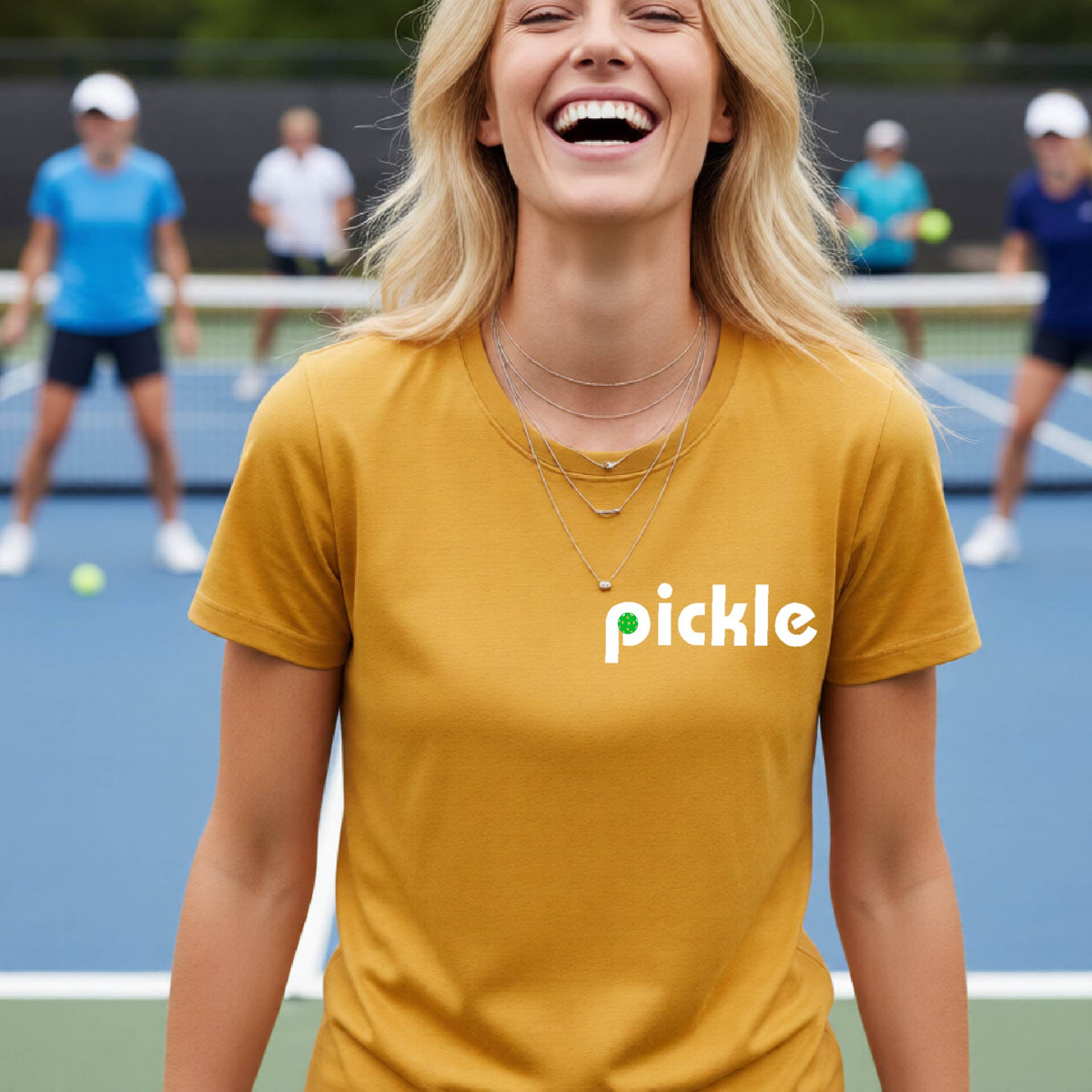 A woman wearing a gold short-sleeve t-shirt with a white graphic of ‘PICKLE’ with a green pickleball integrated into the letter P printed on the front, in front of a pickleball court. 