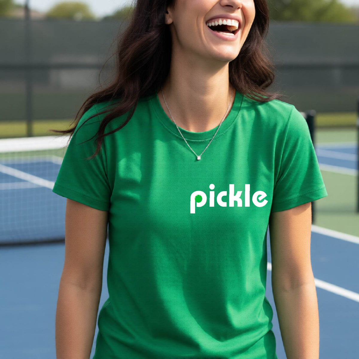 A woman wearing a green short-sleeve t-shirt with a white graphic of ‘PICKLE’ with a green pickleball integrated into the letter P printed on the front, in front of a pickleball court. 