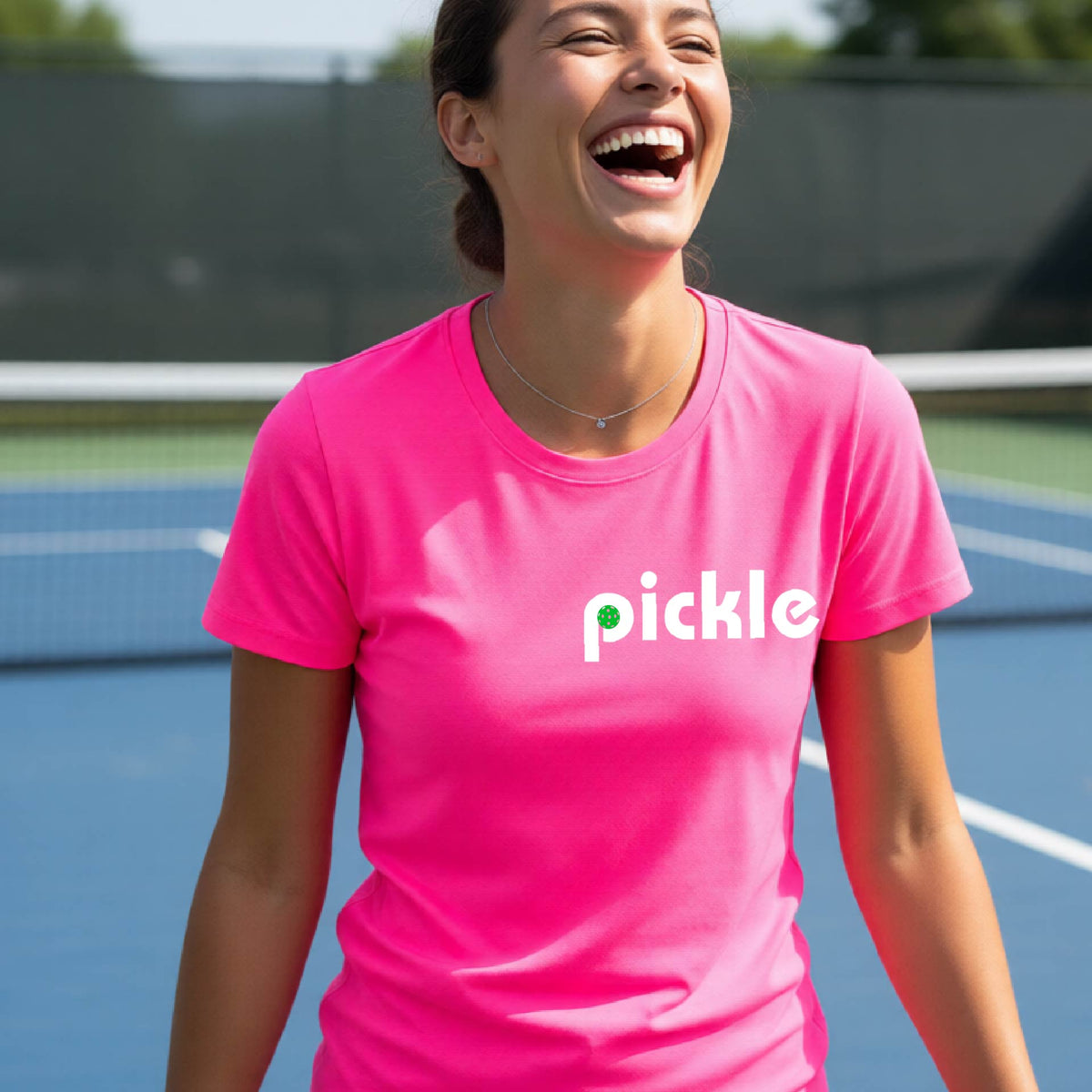 A woman wearing a pink short-sleeve t-shirt with a white graphic of ‘PICKLE’ with a green pickleball integrated into the letter P printed on the front, in front of a pickleball court. 