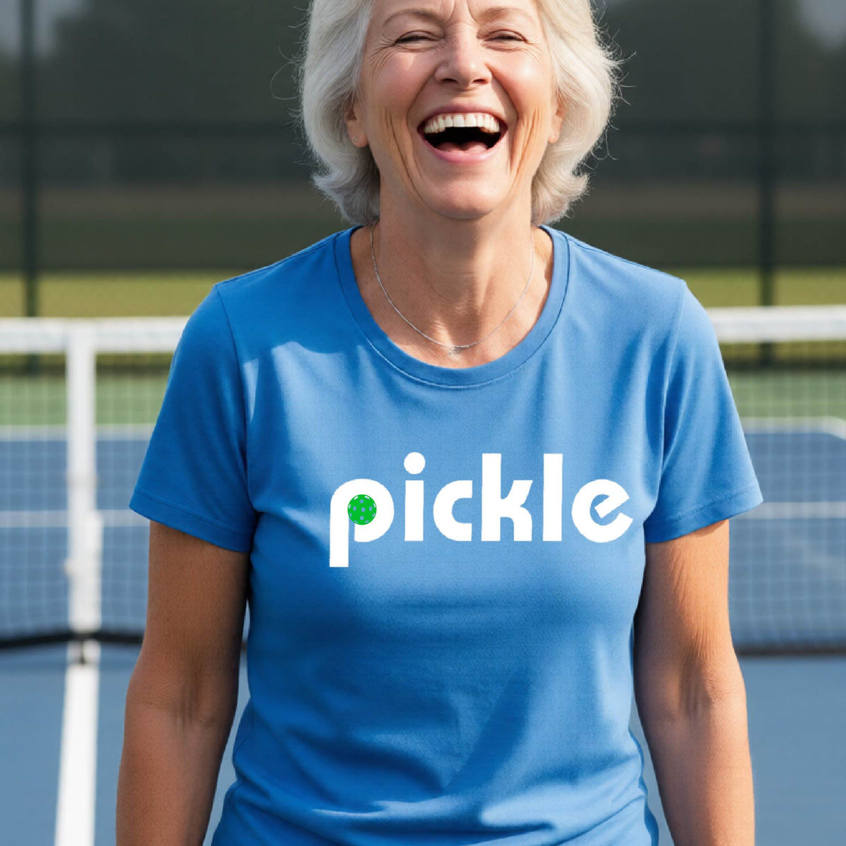 A woman wearing a blue short-sleeve t-shirt with a white graphic of ‘PICKLE’ with a green pickleball integrated into the letter P printed on the front, in front of a pickleball court. 