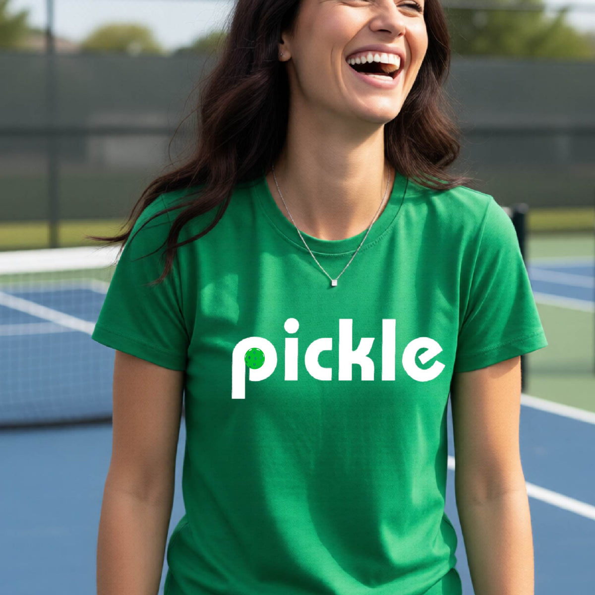 A woman wearing a green short-sleeve t-shirt with a white graphic of ‘PICKLE’ with a green pickleball integrated into the letter P printed on the front, in front of a pickleball court. 