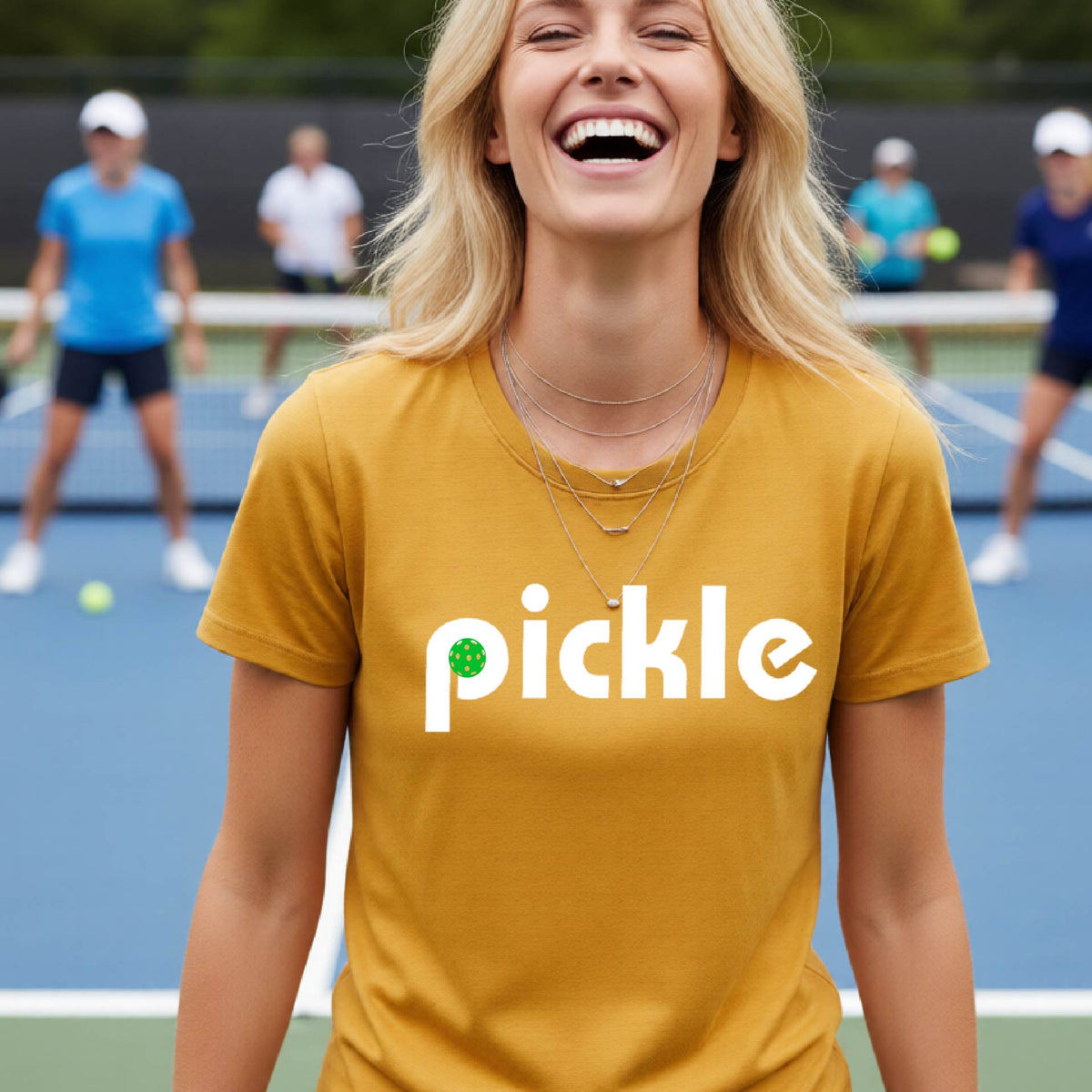 A woman wearing a gold short-sleeve t-shirt with a white graphic of ‘PICKLE’ with a green pickleball integrated into the letter P printed on the front, in front of a pickleball court. 