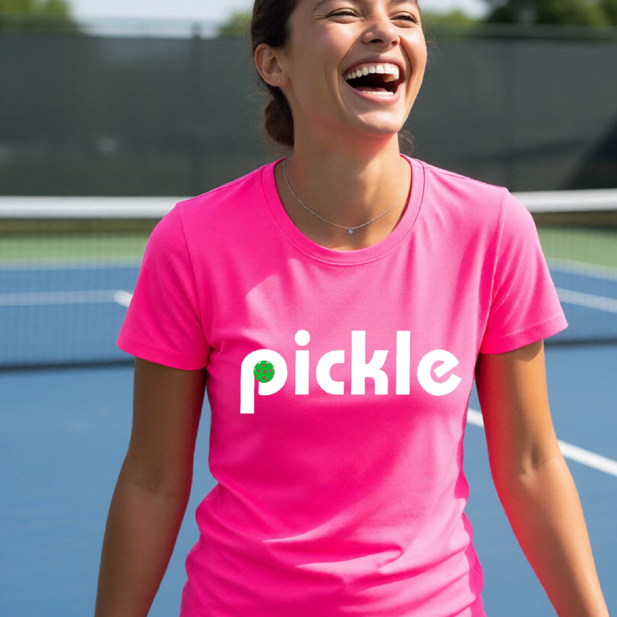A woman wearing a pink short-sleeve t-shirt with a white graphic of ‘PICKLE’ with a green pickleball integrated into the letter P printed on the front, in front of a pickleball court. 
