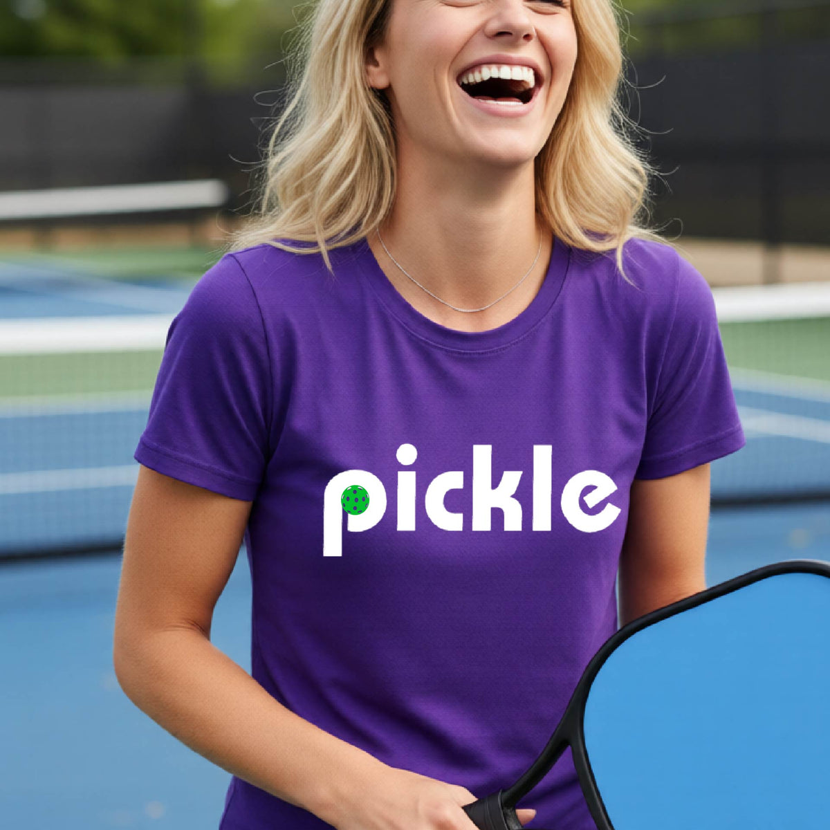 A woman wearing a purple short-sleeve t-shirt with a white graphic of ‘PICKLE’ with a green pickleball integrated into the letter P printed on the front, in front of a pickleball court. 