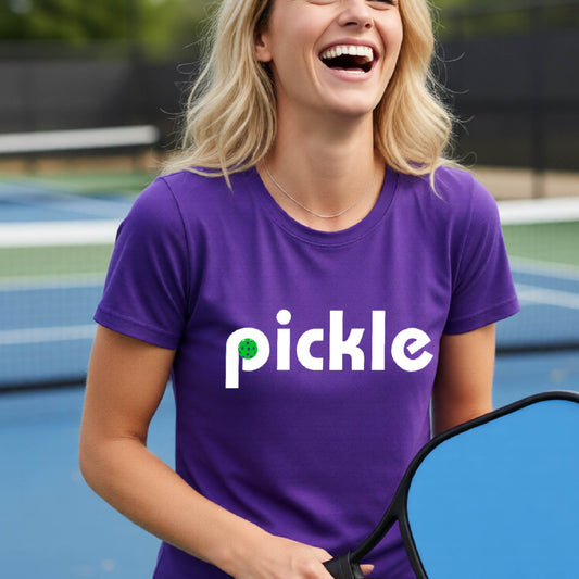 A woman wearing a purple short-sleeve t-shirt with a white graphic of ‘PICKLE’ with a green pickleball integrated into the letter P printed on the front, in front of a pickleball court. 