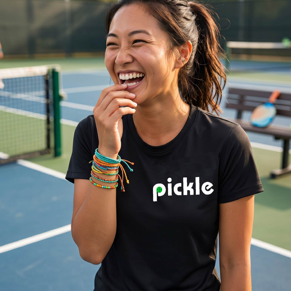 A woman wearing a black short-sleeve t-shirt with a white graphic of ‘PICKLE’ with a green pickleball integrated into the letter P printed on the front, in front of a pickleball court. 